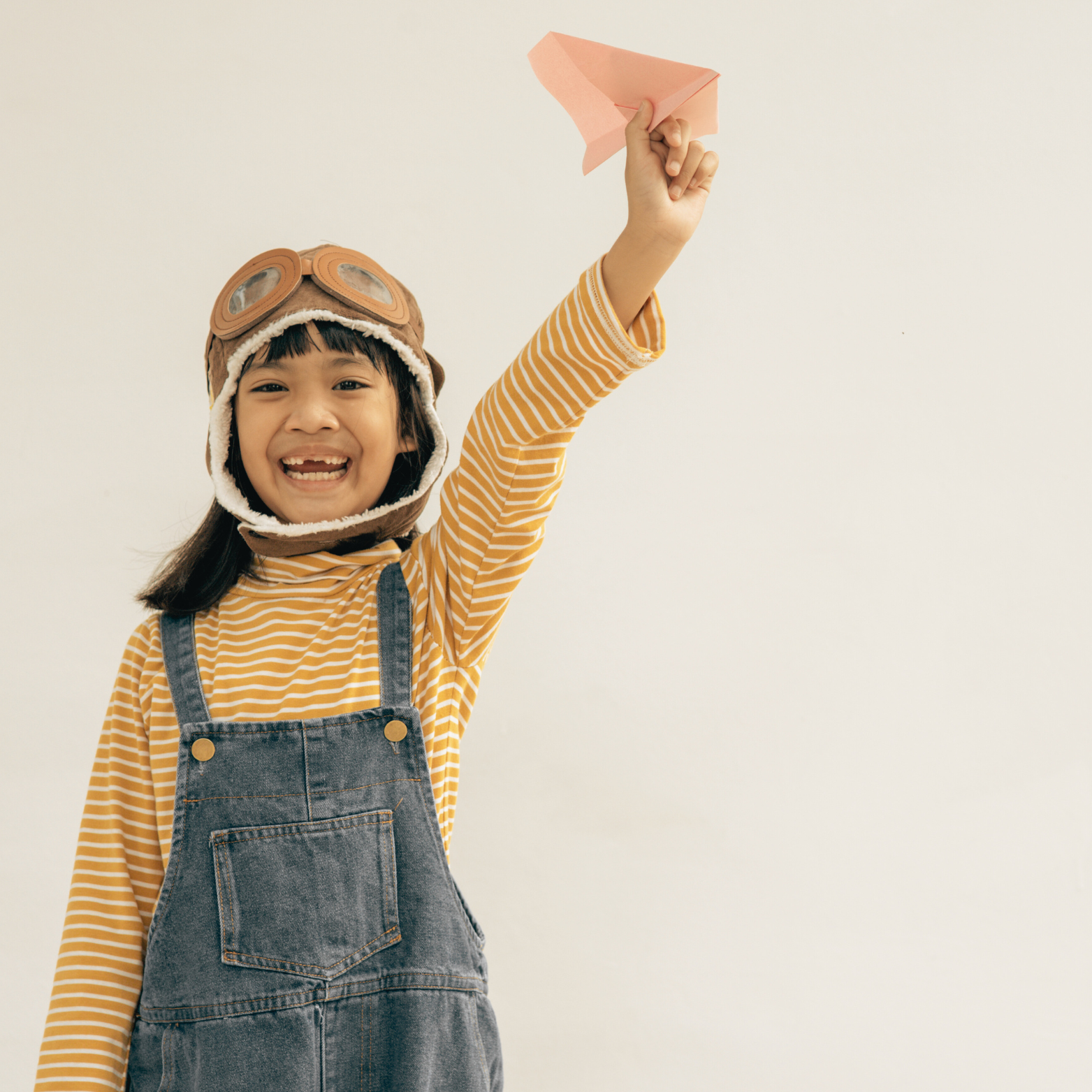 child wearing a yellow striped shirt and overalls getting ready to fly a paper airplane